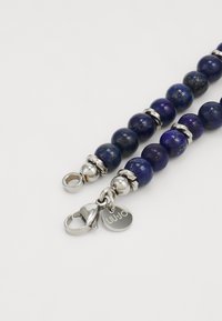 Perlenarmband mit tiefblauen Lapislazuli-Kugeln, durchsetzt mit silberfarbigen Akzenten und einem Verschluss, der ein Markenschild trägt.