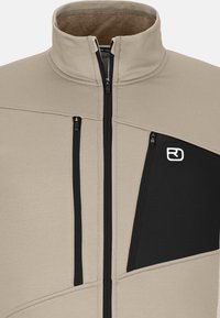 Beige Zip-Jacke mit schwarzen Akzenten, strukturiertem Material, hohem Kragen und zwei Reißverschluss-Brusttaschen, mit einem auffälligen Logo.