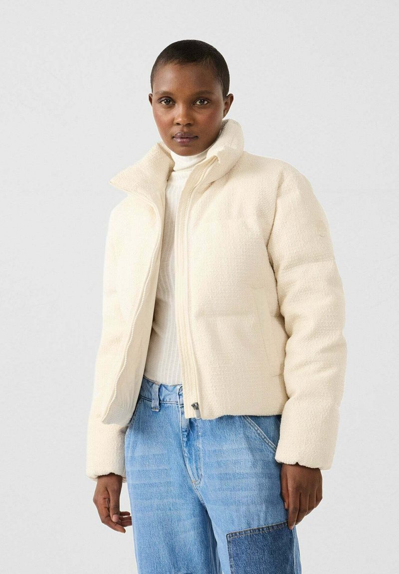 Chaquetón acolchado color crema con un cuello alto, tela texturizada, cremallera frontal y bolsillos; combinado con un suéter blanco de canalé y jeans azules.