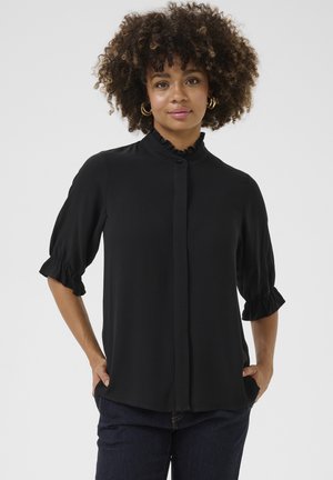 Schwarze Bluse mit hohem, gerafftem Kragen und Puffärmeln. Glattes Material mit Knopfverschluss und gerafften Manschetten. Kombiniert mit dunklen Jeans.