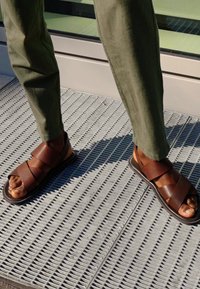 Braune Ledersandalen mit mehreren überkreuzten Riemen, flachen Sohlen und freiliegenden Zehen; kombiniert mit olivgrünen Hosen auf einer karierten Oberfläche.