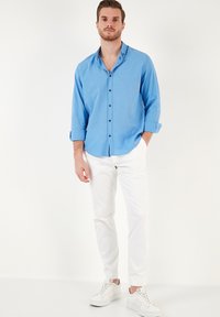 Camisa de botón azul claro con mangas remangadas, botones negros, combinada con pantalones ajustados blancos y zapatillas blancas, sobre un fondo liso.