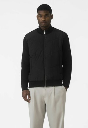 Jachetă neagră cu fermoar frontal, partea superioară matlasată, manșete și tiv canelate, cu buzunare laterale, purtată peste pantaloni deschiși la culoare.