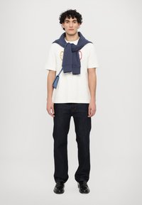 Bela majica s barvnimi vezeninami, mornarsko modra jopica zavezana okrog vratu, temno jean hlače in črne čevlje.