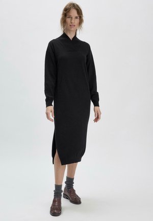 Abito nero lavorato a maglia con maniche lunghe, colletto alto, fessure laterali e una vestibilità rilassata. Indossato con calzini grigi e scarpe marroni.