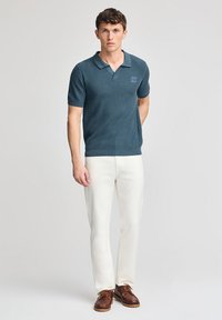 Polo de canalé en azul oscuro con mangas cortas, que presenta un parche de logo en contraste, combinado con pantalones blancos y zapatos marrones con cordones.