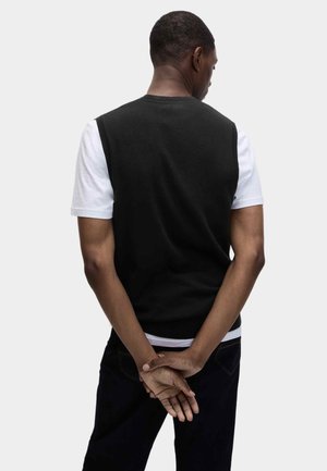 Mann steht mit dem Rücken zur Kamera, trägt schwarzen ärmellosen Pullover über weißem kurzärmligem Hemd, die Hände hinter dem Rücken verschränkt.