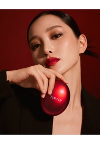 Rood compact met glanzende afwerking, met de merknaam "TIRTIR" in het wit. Model toont het met een neutrale manicure tegen een donkere achtergrond.