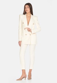Blazer bianco doppiopetto con bottoni dorati, abbinato a pantaloni bianchi aderenti e décolleté nude, che mostrano una vestibilità su misura.