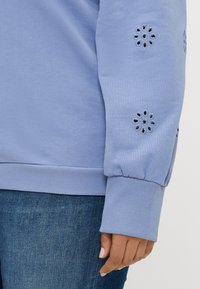 Ανοιχτό μπλε φούτερ με ριγέ μανίκια, διακοσμημένο με λουλουδάτα στοιχεία από δαντέλα στα μανίκια. Συνδυάζεται με σκούρα τζιν παντελόνια.