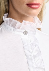 Bela bluza z volanastim ovratnikom, s srebrnim gumbom v obliki cveta. Tkanina se zdi mehka in gladka ter ima rahlo sijaj.