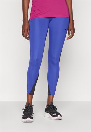 Leggings atléticos azuis com um ajuste justo, apresentando detalhes pretos nos tornozelos e um acabamento suave e texturizado. Usados com ténis pretos.