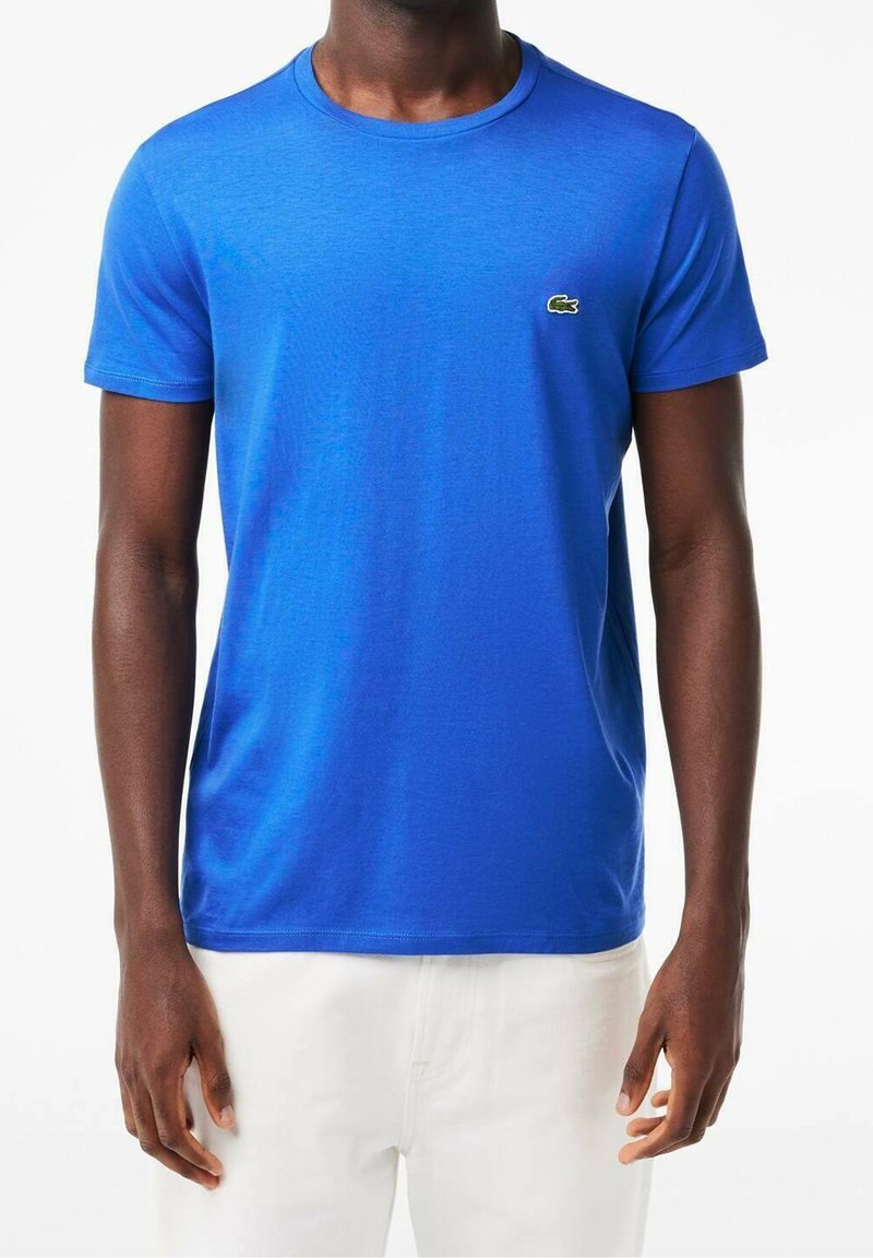 Homme portant un t-shirt bleu vif à manches courtes avec un petit logo crocodile vert sur la poitrine, associé à un pantalon blanc, debout devant un fond blanc.