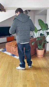 Šedá péřová bunda s kapucí, modré džínové kalhoty a bílé tenisky. Dřevěná podlaha a hnědá pohovka s rostlinou v pozadí.