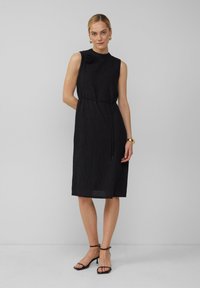Robe noire sans manches avec une finition texturée, dotée d'un nœud sur le côté et d'un accent floral à l'épaule. Portée avec des talons noirs à lanières.