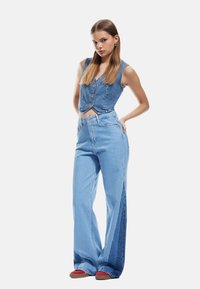 Chaleco de mezclilla cortado con botones, combinado con jeans de pierna ancha de tiro alto que cuentan con paneles laterales en contraste. Color azul claro en todo.