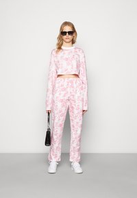 Sweatshirt cropped com padrão tie-dye rosa e calças combinando feitas de um tecido leve e macio. A modelo usa óculos de sol pretos e segura uma mala preta.
