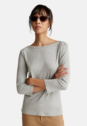 Camicetta grigia a maniche lunghe con scollatura ampia e maniche a tre quarti, realizzata in tessuto morbido. Modello che indossa occhiali da sole, con le braccia incrociate.