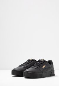 Paire de baskets noires Puma avec semelles nervurées, design à lacets et logo Puma doré sur les côtés et la languette, sur fond blanc.