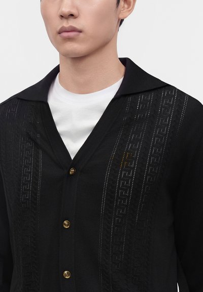 Uomo che indossa un cardigan nero con bottoni e righe verticali decorate sopra una t-shirt bianca girocollo, mostrando collo e parte superiore del petto.