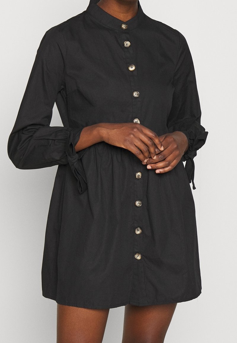 Vestito nero abbottonato realizzato in cotone, caratterizzato da un colletto alto, maniche lunghe con dettagli in una cravatta e una vita arricciata. Bottoni in argento decorano la parte anteriore.
