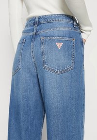 Jasnoniebieskie dżinsy z wysokim stanem, z dwoma tylnymi kieszeniami i małym czerwonym logo w kształcie trójkąta na prawej kieszeni. Teksturowana tkanina.