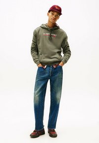 Felpa verde con logo "Tommy Jeans", jeans di denim blu e sneakers rosse/nere. L'outfit presenta una vestibilità rilassata e un design casual.
