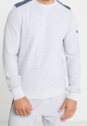 Man draagt lichtgrijze trui met donkergrijze schouderstukken en bijpassende joggingbroek tegen een effen achtergrond.