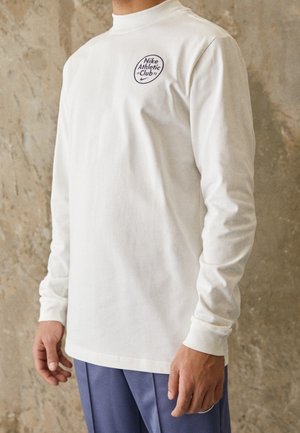Homme portant un t-shirt blanc à manches longues Nike Athletic Club avec logo sur la poitrine et un pantalon bleu, debout contre un mur texturé.