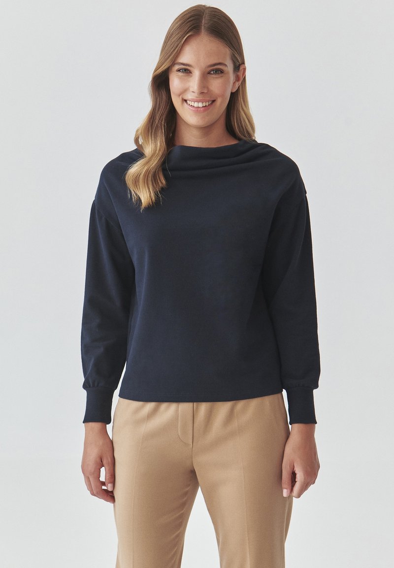 Navy Sweatshirt mit Kapuze und langen Ärmeln, gerippten Bündchen und lockerer Passform, kombiniert mit maßgeschneiderten beigen Hosen. Glatte Stofftextur.