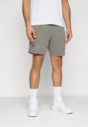 Man draagt olijfgroene Los Angeles Lakers-short, witte sportsokken, witte hoge sneakers en een wit shirt, staand tegen een effen achtergrond.