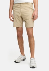 Beige bomullsshorts med rett snitt, frontlommer og beltehemper, paret med hvite og grå joggesko.
