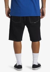 Shorts de mezclilla negros con costuras blancas, dos bolsillos traseros y un ajuste relajado. Se llevan con zapatillas negras y calcetines blancos.