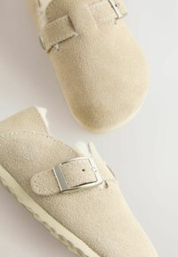 Béžové semišové slip-on topánky s kovovou prackou, mäkkou podšívkou a textúrovanou gumovou podrážkou, s okrúhlym dizajnom špičky.