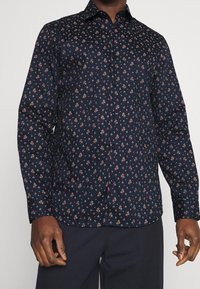 Marinblå skjorta med knappar och små blommönster i rött och vitt. Har en klassisk krage och långa ärmar. Slät tygtextur.