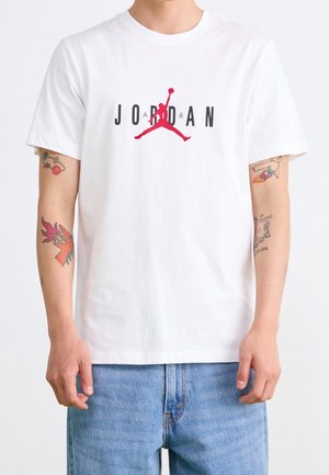 Mand iført hvid Jordan T-shirt med rødt silhuetlogo og blå jeans, der viser farverige tatoveringer på begge underarme.