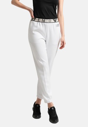 Mujer con pantalones blancos EA7 con cintura elástica con marca, camiseta negra y zapatillas negras, de pie frente a un fondo liso.