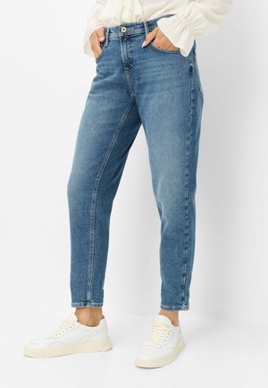 Person, die blaue Slim-Fit-Jeans trägt, mit den Händen in den Taschen und weißen Sneakern vor weißem Hintergrund steht.