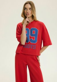 Młoda kobieta w czerwonym stroju z dużą niebieską cyfrą „39” na koszulce zakłada palec na usta w geście ciszy na jednolitym tle.