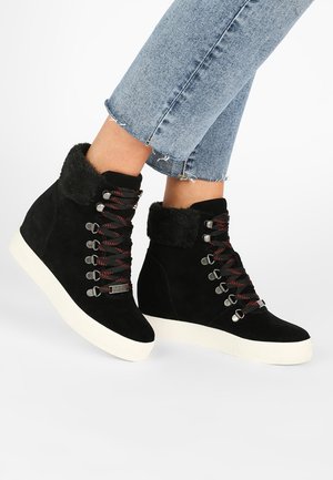Pieds portant des baskets compensées noires avec des lacets rouges et noirs et une bordure en fourrure, associées à un jean court bleu clair.