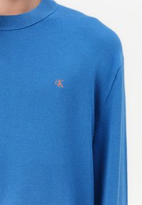 Maglione blu a maglia con collo rotondo a coste e maniche lunghe. Presenta un piccolo logo arancione "CK" sul petto. Tessuto morbido.