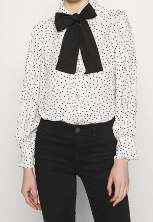 Persoon draagt een witte blouse met lange mouwen met zwarte stippen, een grote zwarte strikdas en zwarte broek met hoge taille.