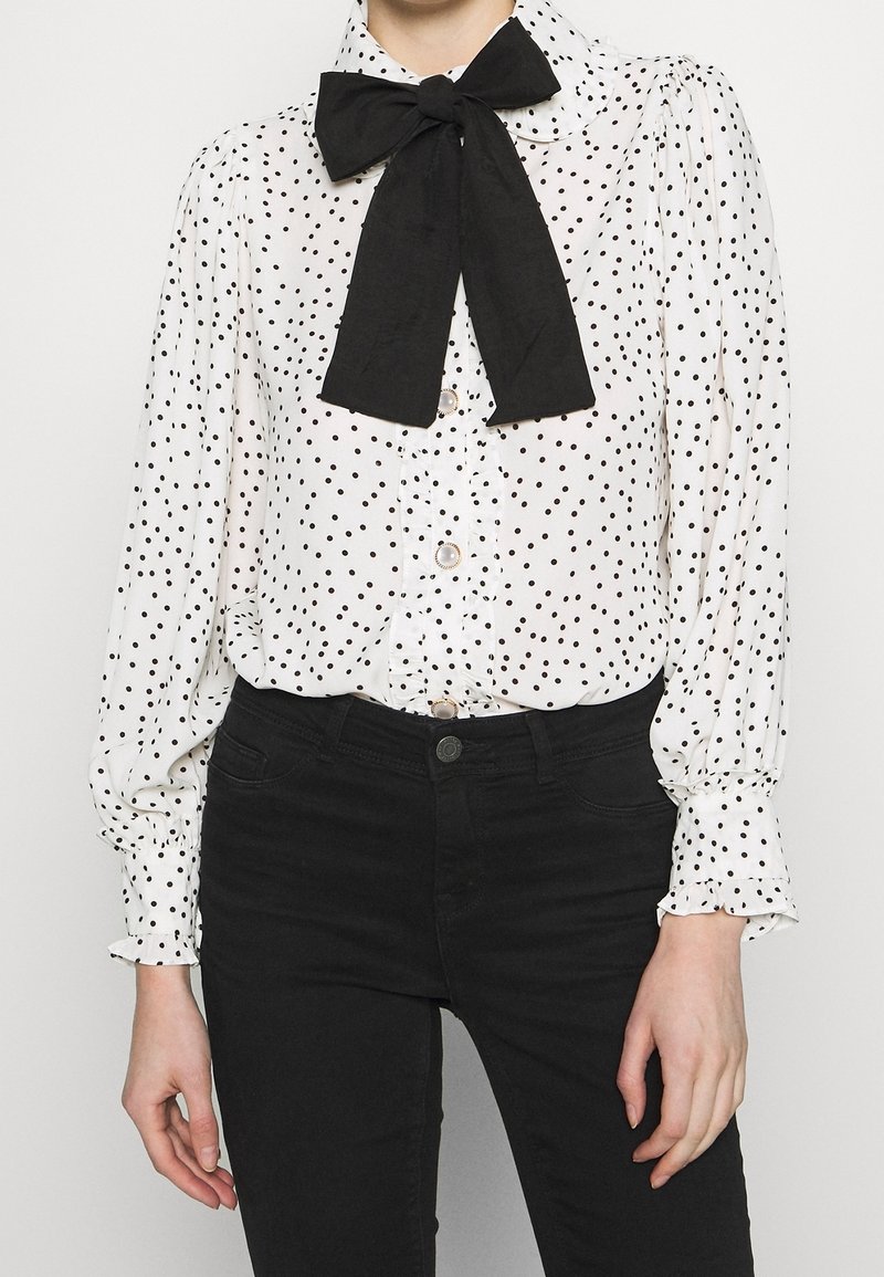 Personne portant un chemisier blanc à manches longues avec des pois noirs, un grand nœud papillon noir et un pantalon noir taille haute.