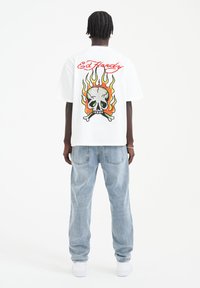 T-shirt bianco a maniche corte con una grafica colorata di teschio e fiamme sulla schiena, abbinato a jeans blu chiaro a vestibilità ampia.