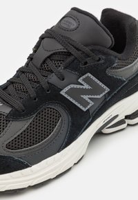 Baskets de sport noires avec des panneaux en mesh et en daim, présentant un logo "N" argenté bien visible, des lacets texturés et une semelle en caoutchouc blanc.