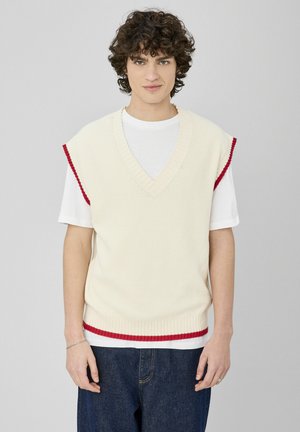 Gilet en maille couleur crème avec un col en V ; présente des accents rouges aux ouvertures des bras et à l'ourlet. Porté sur un T-shirt blanc et associé à un denim foncé.