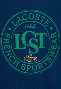 Sudadera azul marino con un logotipo circular verde que dice "Lacoste Paris" y "French Sportswear", además de un detalle bordado de cocodrilo.