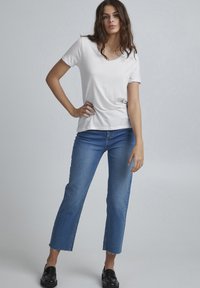 T-shirt bianco a maniche corte con scollo a V, abbinato a jeans blu cropped con orli sfilacciati e vestibilità slim. Scarpe nere slip-on completano il look.