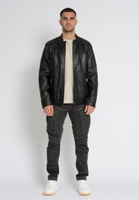 Schwarze Lederjacke mit Reißverschlusstaschen, schlanker Schnitt. Kombiniert mit einem neutralen Hemd und dunklen Cargo-Hosen. Weiße Sneaker runden das Outfit ab.