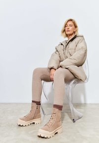 Beige gewatteerde jas met een hoge kraag, geribde tan leggings en tan combat boots met dikke zolen, zittend op een transparante stoel tegen een witte muur.